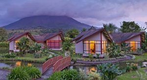 Tres cabañas con luces cálidas, un exuberante jardín, un estanque y un fondo de montañas crean un entorno romántico bajo el cielo nublado del atardecer, perfecto para una escapada en pareja con "Viaje por Costa Rica en pareja". - ForTwo - Planes para parejas