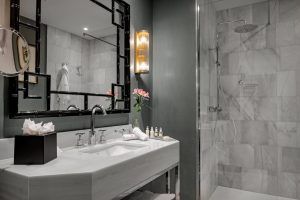 El Hotel NH Collection Colón cuenta con un moderno baño con lavabo de mármol, espejo retroiluminado, jarrón de flores y ducha a ras de suelo con cabezal de lluvia, ofreciendo lujo y confort en el corazón de Valencia. - ForTwo - Planes para parejas