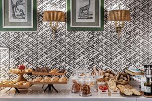 Los productos de panadería están elegantemente dispuestos en un mostrador frente a un papel tapiz estampado, complementados con arte enmarcado y dos elegantes lámparas de pared encima, creando una configuración encantadora que recuerda la elegancia atemporal del Hotel NH Collection Colón en Valencia. - ForTwo - Planes para parejas
