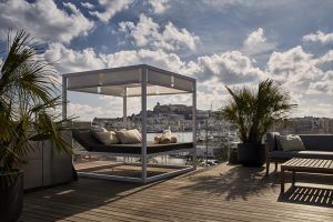 El Hotel Sir Joan de Ibiza cuenta con un patio en la azotea con un diván, exuberantes plantas en macetas y una impresionante vista del puerto de Ibiza y del horizonte de la ciudad bajo un cielo parcialmente nublado. - ForTwo - Planes para parejas
