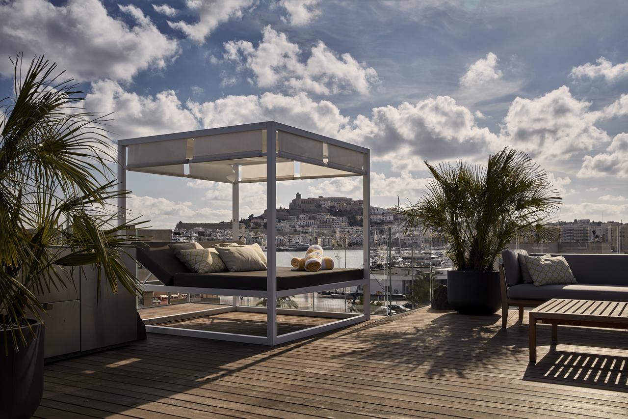 El Hotel Sir Joan de Ibiza cuenta con un patio en la azotea con un diván, exuberantes plantas en macetas y una impresionante vista del puerto de Ibiza y del horizonte de la ciudad bajo un cielo parcialmente nublado. - ForTwo - Planes para parejas