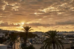 Una puesta de sol sobre un puerto deportivo de Ibiza muestra siluetas de barcos, palmeras y un avión en un cielo nublado y naranja cerca del Hotel Sir Joan (Ibiza). - ForTwo - Planes para parejas