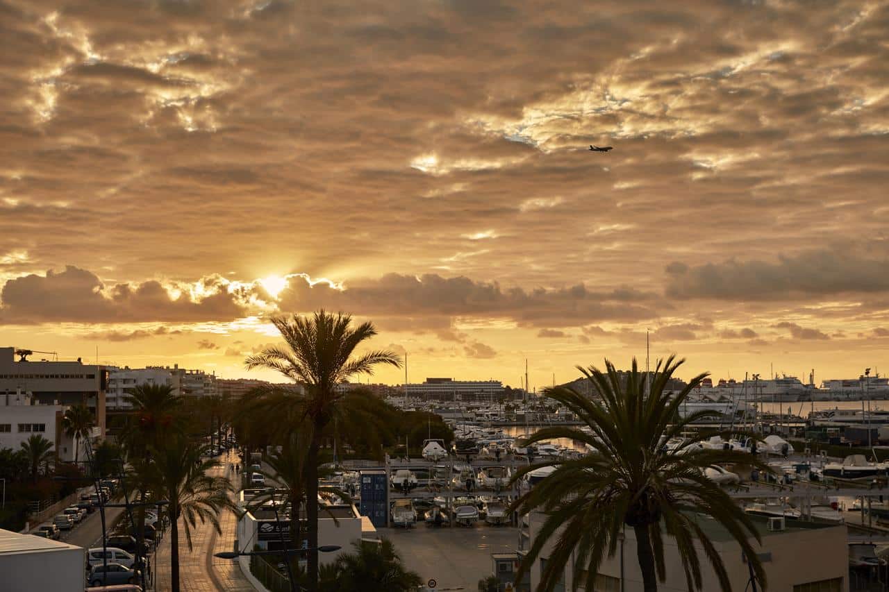 Una puesta de sol sobre un puerto deportivo de Ibiza muestra siluetas de barcos, palmeras y un avión en un cielo nublado y naranja cerca del Hotel Sir Joan (Ibiza). - ForTwo - Planes para parejas