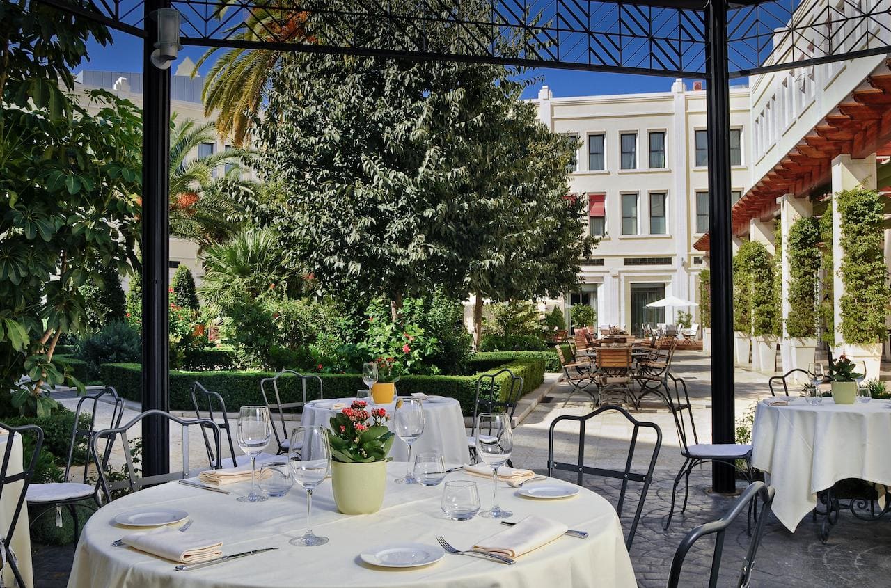 El patio del restaurante al aire libre, ubicado en el corazón de Valencia, en el Hotel The Westin, cuenta con mesas redondas con manteles blancos y macetas con plantas. Rodeado de exuberante vegetación y encantadores edificios, ofrece un remanso de paz. - ForTwo - Planes para parejas