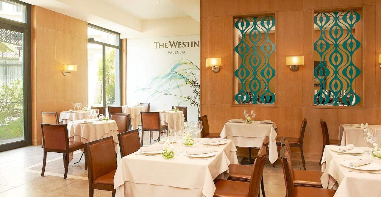 El Hotel The Westin (Valencia) cuenta con un elegante interior de restaurante con mesas cuidadosamente dispuestas, amplios ventanales, paneles de pared decorativos e iluminación suave. - ForTwo - Planes para parejas