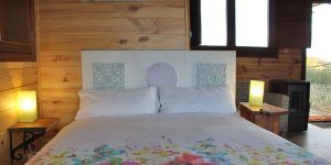Un dormitorio acogedor con paredes de madera, un edredón floral en la cama, iluminación suave y una ventana con vista a la vegetación, ideal para quienes sueñan con Cabaña Aire en Dormir en los árboles en la serena campiña de Cáceres. - ForTwo - Planes para parejas