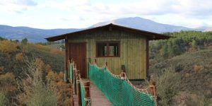 Cabaña Aire en Dormir en los árboles, una cabaña de madera en Cáceres, ofrece una experiencia única. Encaramada en una colina y accesible mediante un puente colgante, está rodeada de árboles y montañas bajo un cielo parcialmente nublado. - ForTwo - Planes para parejas
