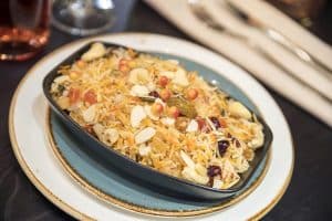 En Sabores de India en Madrid, la Oferta degustación presenta un tazón de biryani adornado con almendras fileteadas, pasas y guarniciones en un plato en capas sobre un fondo borroso. - ForTwo - Planes para parejas