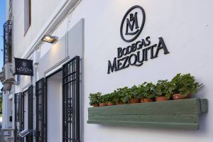 El exterior de Bodegas Mezquita en Córdoba luce su logo y una encantadora jardinera con exuberante vegetación contra la pared blanca, invitándote a probar la "Oferta: cena para dos en Bodegas Mezquita, menú especial Mezquita (Córdoba). - ForTwo - Planes para parejas