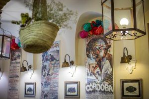 Una pared interior vibrante, con carteles y decoración coloridos, incluye un encantador cartel con temática de Córdoba y deliciosas piezas colgantes, que evocan el romance de una "Oferta: cena para dos en Bodegas Mezquita, menú especial Mezquita (Córdoba). - ForTwo - Planes para parejas