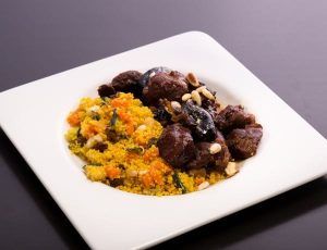 Un plato cuadrado con vibrante cuscús y trozos de carne, aderezado con nueces, resalta la "Oferta: cena para dos en Bodegas Mezquita, menú especial Mezquita (Córdoba)" sobre un fondo oscuro. - ForTwo - Planes para parejas