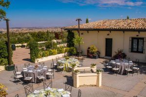 El patio exterior de esta villa rústica en Madrid cuenta con mesas preparadas, ideales para relajarse. Rodeado de exuberante vegetación y un paisaje lejano bajo un cielo despejado, es un lugar idílico para disfrutar de la experiencia de una cata de vinos en la bodega El Regajal (Madrid). - ForTwo - Planes para parejas