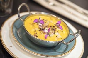 Un plato de sopa amarilla, enriquecida con la Oferta degustación Sabores de India, adornado con flores moradas en un recipiente de metal sobre platos apilados, captura la esencia del Restaurante Bangalore. - ForTwo - Planes para parejas