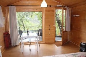La acogedora cabaña de madera, similar a una Cabaña Musgo en Dormir en los Árboles (Cáceres), incluye mesa, sillas y una botella de vino. A través de las puertas de cristal, disfrute de las tranquilas vistas a los árboles que recuerdan a las de los alrededores de Cáceres. - ForTwo - Planes para parejas