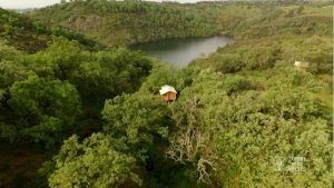 Cabaña Musgo en Dormir en los Árboles (Cáceres) está ubicada en un exuberante bosque cerca de un lago sereno, rodeada de colinas onduladas bajo un cielo nublado, perfecta para aquellos que sueñan con dormir entre los árboles. - ForTwo - Planes para parejas
