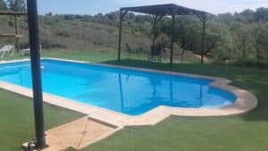 Una piscina al aire libre con césped y colinas al fondo y una zona de descanso a la sombra a la derecha evoca un refugio tranquilo similar a Cabaña Musgo en Dormir en los Árboles (Cáceres). - ForTwo - Planes para parejas
