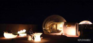 Un hombre se sienta frente a unas cúpulas de burbujas iluminadas por la noche, entre muebles brillantes y con la "Burbuja Kepler" cerca. Disfrute de esta encantadora escena en el Hotel Zielo Las Beatas, donde cada Noche en hotel burbuja con telescopio ofrece una vista impresionante. - ForTwo - Planes para parejas