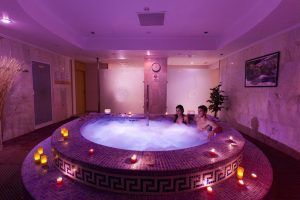 En una sala de spa poco iluminada de Tenerife, una pareja se relaja en un jacuzzi circular rodeado de velas encendidas, disfrutando del Circuito termal de spa - Tratamiento deluxe. - ForTwo - Planes para parejas