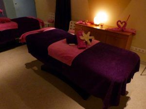 La sala de masajes es un lujoso santuario de spa con una mesa cubierta con toallas de color púrpura y rosa, suavemente iluminada por una lámpara cálida, ideal para disfrutar del Circuito termal de spa - Tratamiento deluxe en Tenerife. - ForTwo - Planes para parejas