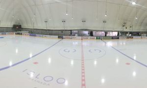 La pista de hockey sobre hielo cubierta y vacía, que recuerda a un iglú helado, presenta vibrantes líneas rojas y azules. Rodeada de anuncios y asientos, invita a los espectadores a disfrutar del espectáculo de "Patinaje sobre hielo para dos en Iglú (Granada)". - ForTwo - Planes para parejas