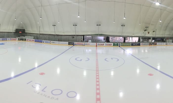 La pista de hockey sobre hielo cubierta y vacía, que recuerda a un iglú helado, presenta vibrantes líneas rojas y azules. Rodeada de anuncios y asientos, invita a los espectadores a disfrutar del espectáculo de "Patinaje sobre hielo para dos en Iglú (Granada)". - ForTwo - Planes para parejas