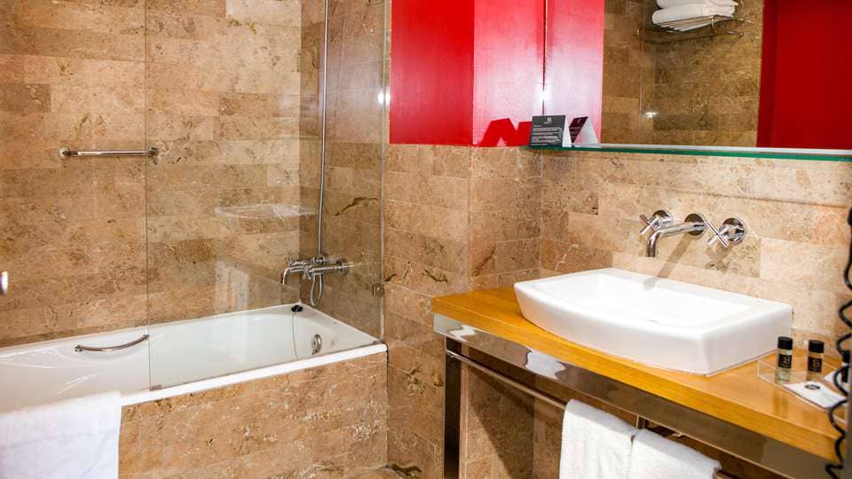 El baño, con azulejos beige, ofrece un escape de lujo similar al de Escapada Hotel Soho Boutique Casa Don Fernando. Un elegante lavabo blanco sobre una encimera de madera y una bañera con paneles de vidrio combinan estilo y funcionalidad para crear un santuario perfecto. - ForTwo - Planes para parejas