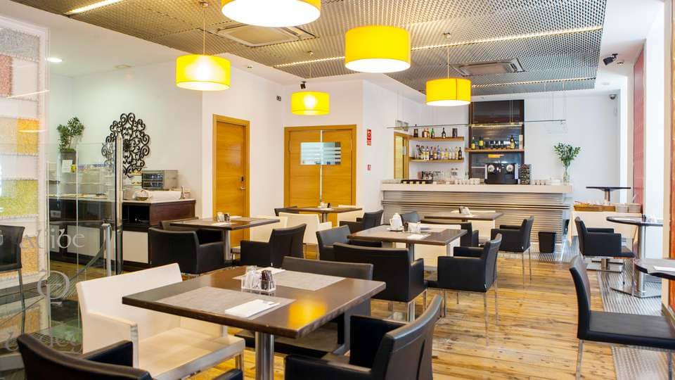 Disfrute de una gastronomía moderna en el centro de Cáceres con una elegante combinación de sillas blancas y negras, mesas de madera y lámparas colgantes amarillas en un elegante bar. El ambiente refleja la elegancia de Soho Boutique Casa Don Fernando. Disfrute de un desayuno en un ambiente que combina el encanto clásico y contemporáneo. - ForTwo - Planes para parejas