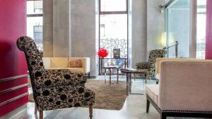 El salón del Hotel Soho Boutique cuenta con sillas estampadas, un sofá beige, una pared roja y una escultura geométrica. Refleja el lujo del Escapada Hotel Soho Boutique Casa Don Fernando en Cáceres. - ForTwo - Planes para parejas