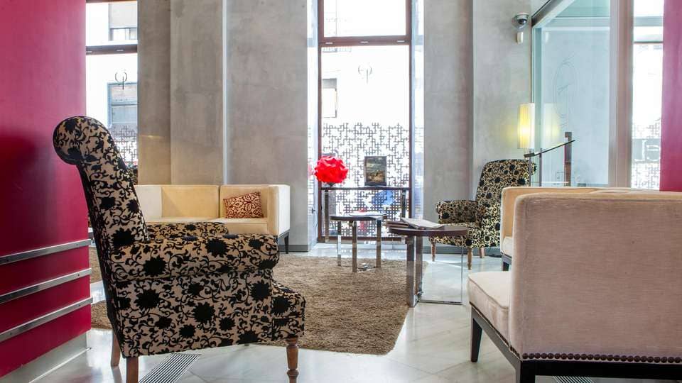 El salón del Hotel Soho Boutique cuenta con sillas estampadas, un sofá beige, una pared roja y una escultura geométrica. Refleja el lujo del Escapada Hotel Soho Boutique Casa Don Fernando en Cáceres. - ForTwo - Planes para parejas