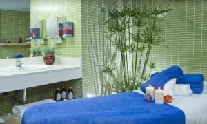Descubra un remanso de paz con la "Sesión de spa en pareja" del Hotel Gala, Tenerife. Disfrute de un ambiente tranquilo con una camilla de masaje con toalla azul, velas encendidas y exuberante vegetación para una relajación total. - ForTwo - Planes para parejas