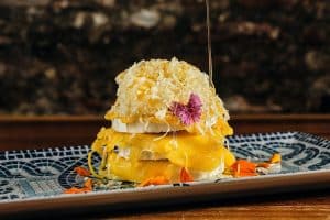 Postre en capas adornado con rodajas de mango, queso rallado y una flor rosa, rociado con almíbar sobre un elegante plato estampado, ideal para una cena romántica en La Tabernilla del Darro en Granada. - ForTwo - Planes para parejas