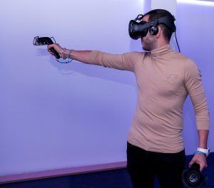 En la sala iluminada de azul de San Sebastián de los Reyes, una persona plenamente inmersa en la Realidad Virtual en Pareja de Oasis VR Experience permanece de pie con un brazo extendido, equipado con un casco de realidad virtual y un controlador. - ForTwo - Planes para parejas