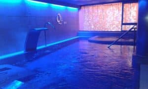 Sumérgete en la tranquilidad de la piscina cubierta de Qi Spa en Bilbao. Disfruta de la relajante iluminación azul, la suave cascada y la exclusiva zona de jacuzzi, todo ello parte de nuestro lujoso "Circuito spa para 2 personas en Qi Spa (Bilbao)", bellamente realzado por una cálida iluminación de fondo. - ForTwo - Planes para parejas