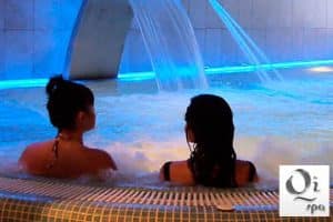 En el reconocido Qi Spa de Bilbao, dos personas se relajan en una piscina de hidromasaje con chorros de agua relajantes e iluminación azul ambiental. El logotipo de Oi Spa se distingue sutilmente en la esquina. Producto: Circuito spa para 2 personas en Qi Spa (Bilbao). - ForTwo - Planes para parejas