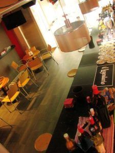 Vista en ángulo de un bar con reminiscencias bilbaínas, con botellas, mesas redondas, sillas y brillantes lámparas colgantes. El animado ambiente invita a disfrutar de una velada con el "Combinado de mojitos y pintxos únicos (Bilbao)". - ForTwo - Planes para parejas