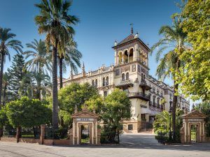 Experimente la máxima elegancia en el Hotel Alfonso XIII en Sevilla, donde la Suite Deluxe combina a la perfección el encanto histórico con la comodidad moderna, todo ello rodeado de elegantes palmeras y una intrincada arquitectura bajo un cielo azul claro. - ForTwo - Planes para parejas