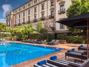 Ubicado junto al histórico Hotel Alfonso XIII de Sevilla, el Máxima elegancia en la Suite Deluxe ofrece una piscina al aire libre con tumbonas y toallas a rayas, a la sombra de sombrillas, con un telón de fondo de ventanas arqueadas y frondosos árboles. - ForTwo - Planes para parejas