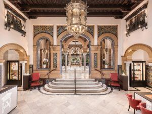 En el elegante vestíbulo del Hotel Alfonso XIII, arcos ornamentados y azulejos intrincados enmarcan una gran lámpara de araña, mientras sillas rojas flanquean los escalones de mármol que conducen a la lujosa Máxima elegancia en la Suite Deluxe en el hotel Alfonso XIII en Sevilla. - ForTwo - Planes para parejas