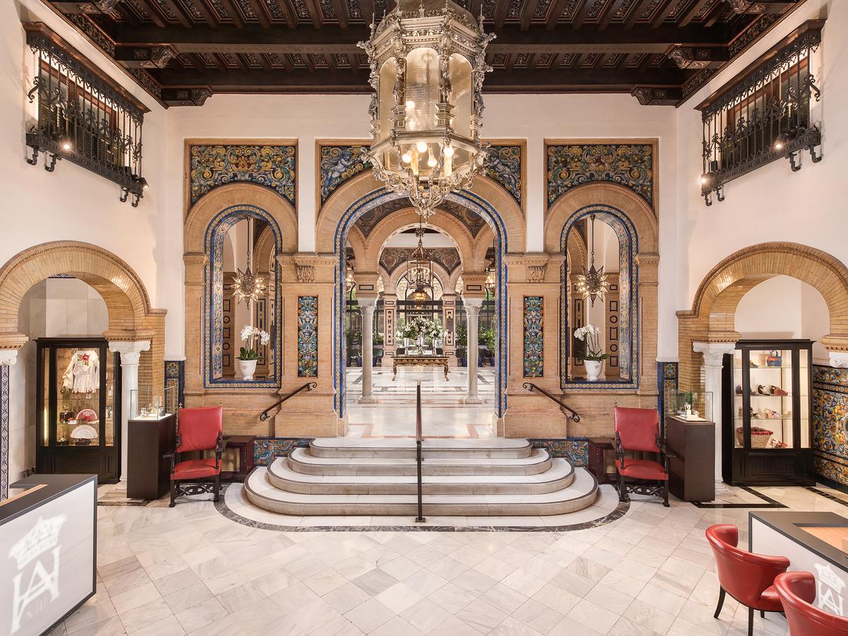 En el elegante vestíbulo del Hotel Alfonso XIII, arcos ornamentados y azulejos intrincados enmarcan una gran lámpara de araña, mientras sillas rojas flanquean los escalones de mármol que conducen a la lujosa Máxima elegancia en la Suite Deluxe en el hotel Alfonso XIII en Sevilla. - ForTwo - Planes para parejas
