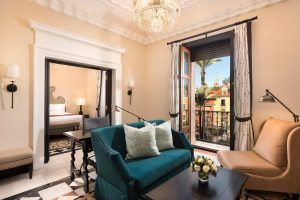 En la Suite Deluxe del Hotel Alfonso XIII, Máxima elegancia ofrece un sofá verde azulado, un sillón y una lámpara de araña con puertas de balcón que se abren a una vista impresionante de los vibrantes edificios de Sevilla, combinando la comodidad moderna con el encanto clásico. - ForTwo - Planes para parejas