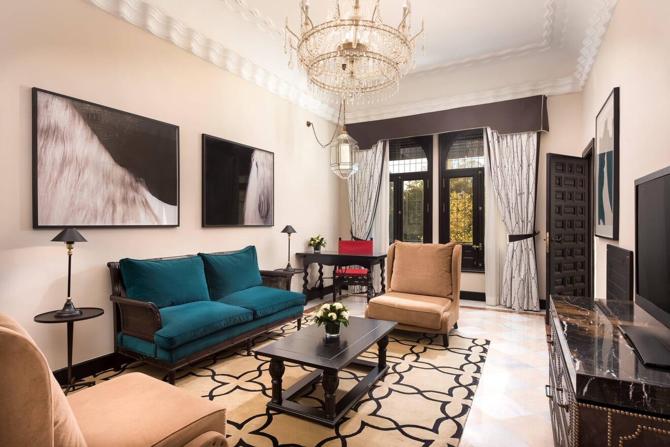 Una sala de estar evoca la máxima elegancia en la Suite Deluxe del Hotel Alfonso XIII, con un sofá azul, sillas marrones, una lámpara de araña y una exquisita obra de arte en la pared. Los amplios ventanales dejan entrar la luz natural, realzando el encanto de la alfombra estampada. - ForTwo - Planes para parejas