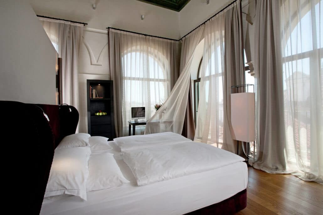 Disfrute de la luminosa habitación del Hotel Palacio de Villapanés con cortinas transparentes, una cama impecablemente tendida y un pequeño escritorio junto a los altos ventanales. Descubra Sevilla con nuestras suites únicas en el Hotel Palacio de Villapanés, que combinan elegancia y confort. - ForTwo - Planes para parejas