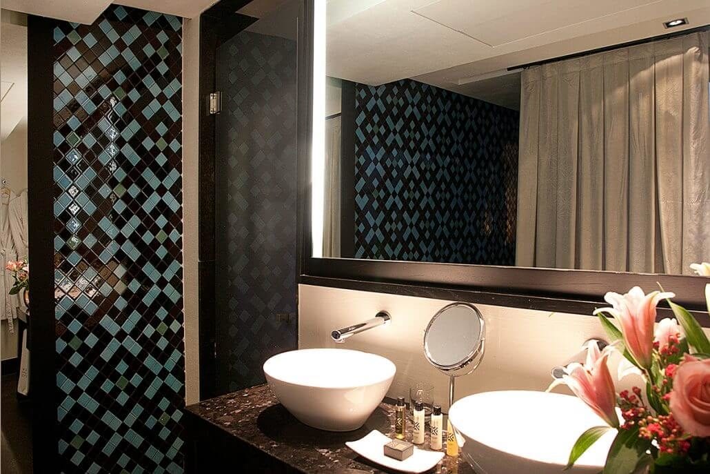 Las Suites únicas del Hotel Palacio De Villapanés en Sevilla cuentan con un moderno baño con azulejos de mosaico y elegantes lavabos, acentuados por un espejo y flores frescas sobre una encimera oscura, capturando la esencia del encanto atemporal de Sevilla. - ForTwo - Planes para parejas