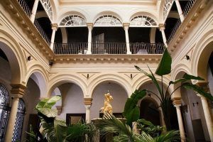 Disfrute del tranquilo encanto del Hotel Palacio de Villapanés en Sevilla, con balcones arqueados, una estatua amarilla y exuberante vegetación bajo un techo alto. Descubra suites únicas que fusionan a la perfección el lujo con la elegancia clásica. - ForTwo - Planes para parejas