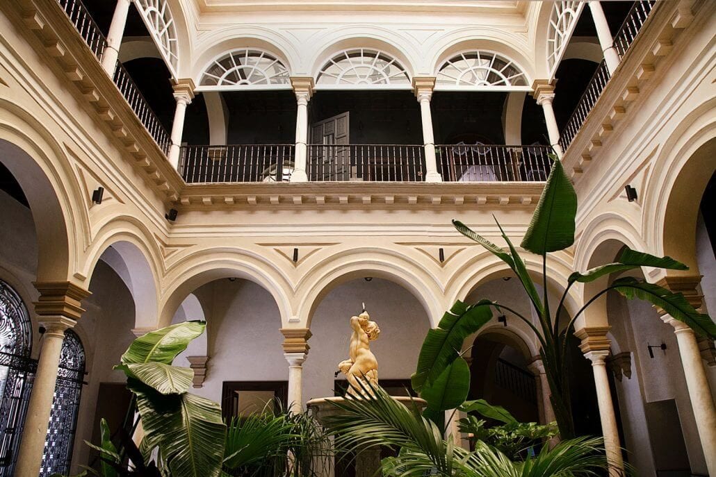Disfrute del tranquilo encanto del Hotel Palacio de Villapanés en Sevilla, con balcones arqueados, una estatua amarilla y exuberante vegetación bajo un techo alto. Descubra suites únicas que fusionan a la perfección el lujo con la elegancia clásica. - ForTwo - Planes para parejas