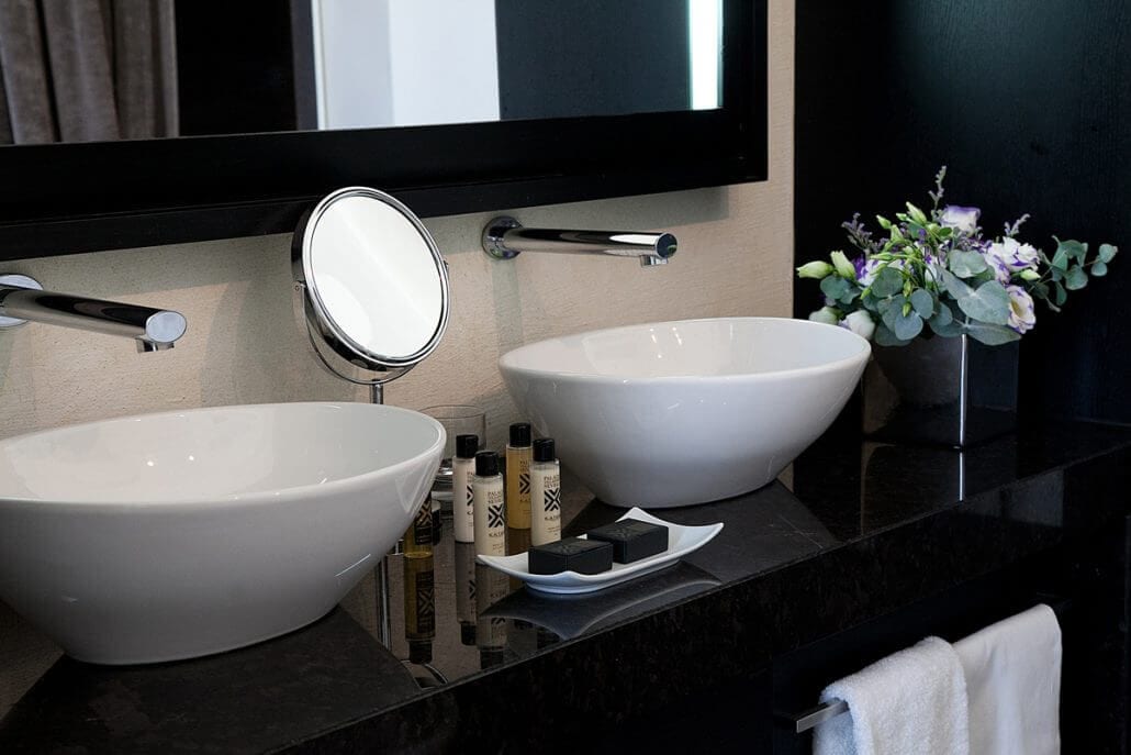 Este moderno baño, que recuerda a las Suites únicas del Hotel Palacio De Villapanés (Sevilla), cuenta con dos lavabos blancos, un espejo redondo, artículos de tocador y un jarrón con flores sobre una elegante encimera negra. - ForTwo - Planes para parejas