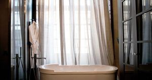 El baño de las Suites únicas del Hotel Palacio De Villapanés, Sevilla, incluye una bañera blanca, cortinas transparentes y una lujosa bata en la pared. - ForTwo - Planes para parejas