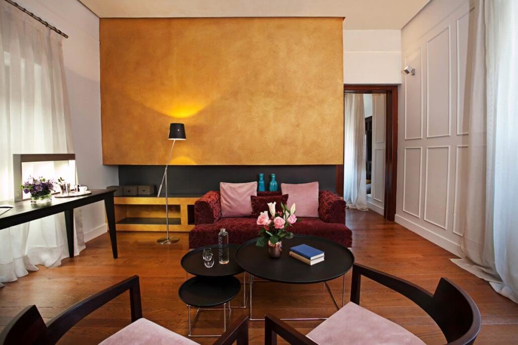 Dentro del Hotel Palacio De Villapanés en Sevilla, encontrará una sala de estar con un sofá rojo, una pared con acento dorado, una mesa redonda negra y flores decorativas, parte de sus suites únicas. - ForTwo - Planes para parejas