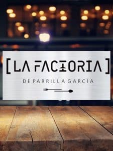 Un cartel que reza "La Factoría de Parrilla García" adorna una mesa de madera, rodeada de luces difusas, invitando a los comensales a saborear la encantadora experiencia culinaria de Tenerife con el "Menú para dos La Factoría de Parrilla García". - ForTwo - Planes para parejas