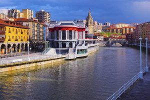 Disfrute de una colorida vista del río con sus edificios y un gran mercado, con un puente bajo un cielo soleado. Capture esta escena en la cima de Artxanda con el funicular o sumérjase en ella con el "Tour guiado + funicular de Artxanda (Bilbao)" para una aventura inolvidable. - ForTwo - Planes para parejas